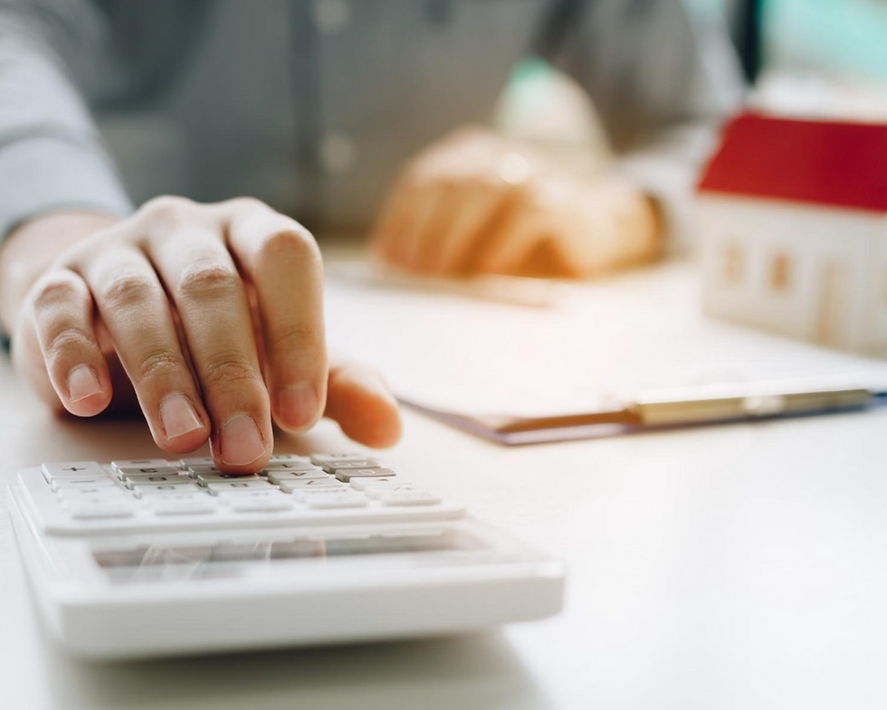 Hand pressing calculator keys on a white desk with a small house model and clipboard nearby, in a softly lit room, suggesting focus.