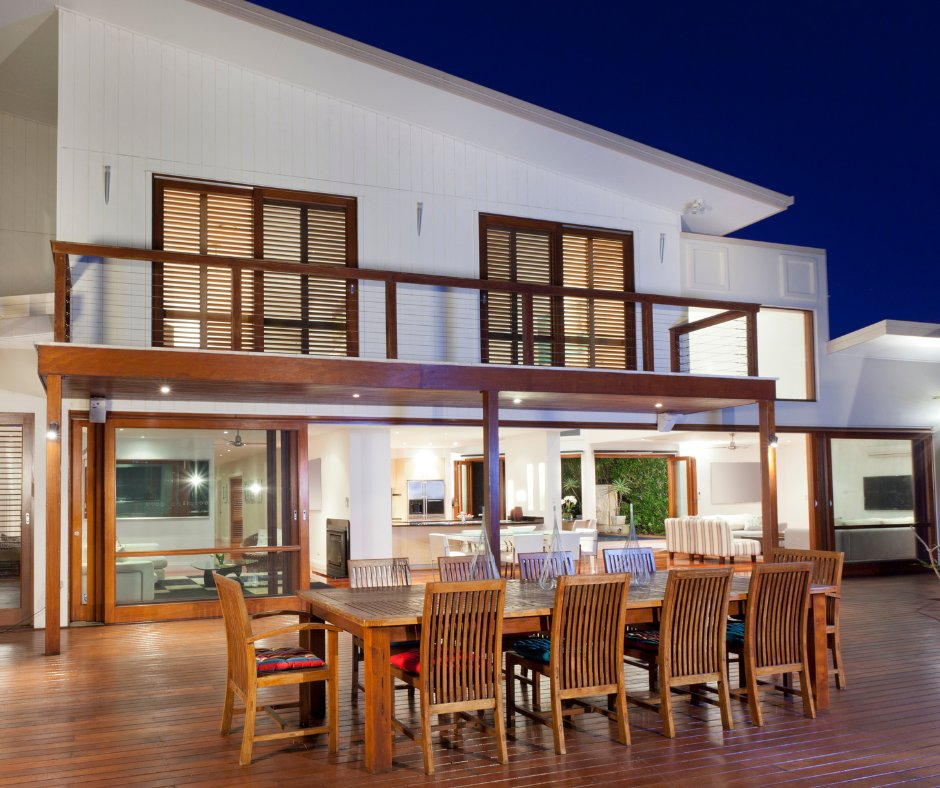 Modern house with wooden deck and dining table, chairs, and large windows. Evening setting with warm indoor lights and dark blue sky.