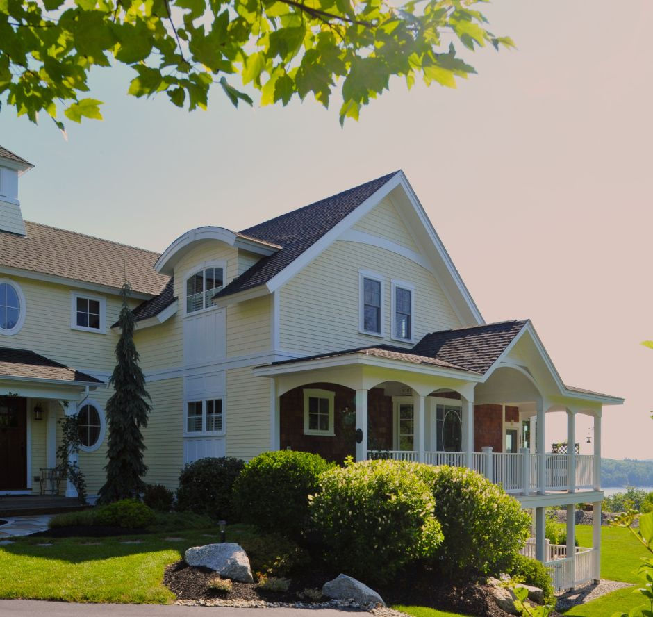A classic, light yellow, custom New England-style home with white trim sits on a sun-lit lakefront hillside in NH.
