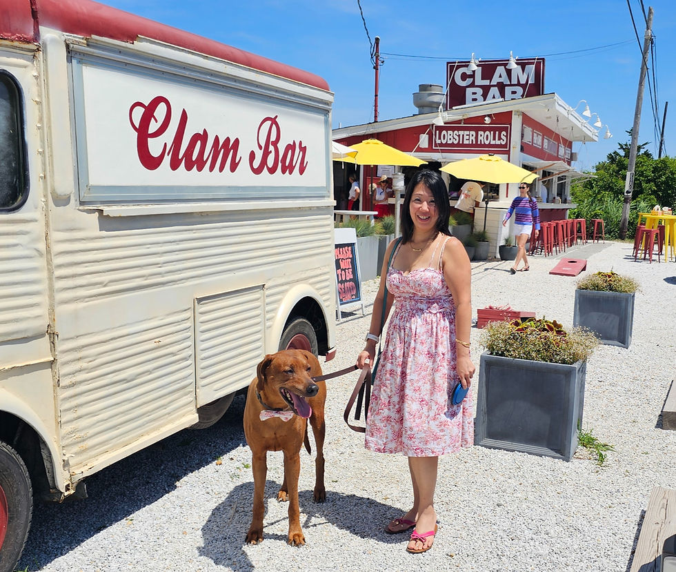 Smiling woman with brown dog outside a Clam Bar seafood shack, with Lobster Rolls sign and yellow umbrellas on a sunny day