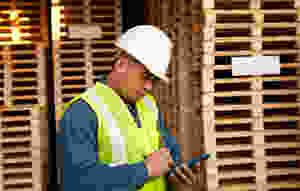 operario fazendo apontamenot eletrônico em seu smartphone, entre palets, com capacete branco e roupa de segurança