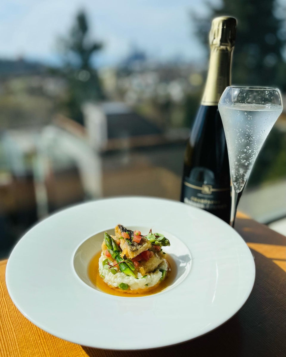 Black cod and asparagus on a rice pilaf with bouillabaisse on a white bowl with a glass and bottle of cava on the right hand side. A seasonal French inspired offering at iconiq.