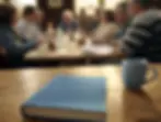 Big book of AA and mug on table in focus, people in casual recovery discussion in blurred sourth florida interior background. Warm, relaxed setting.