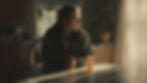 A woman in a dim room sits at a table with a prescription bottle. considering if her MAT status has limited her recovery housing options. Sunlight streams through curtains, creating a contemplative mood.