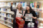 Two people in front of library books are looking at a microfiche that one person is holding up to the light. The other person is holding a newpaper facing the viewer.