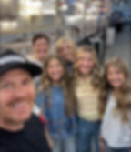 Group selfie of smiling man and four women in a lighting store aisle, with hanging lights, boxes, and a LIVE ELEVATED cap.