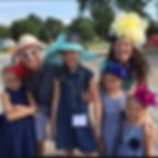Smiling group of adults and girls in blue dresses and oversized colorful hats posing outdoors on a sunny day.
