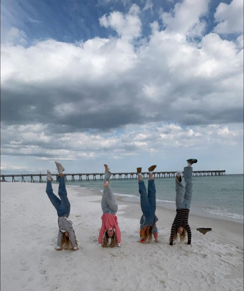 Four girls do handstands on a white beach under cloudy skies, with a long pier stretching into the sea.