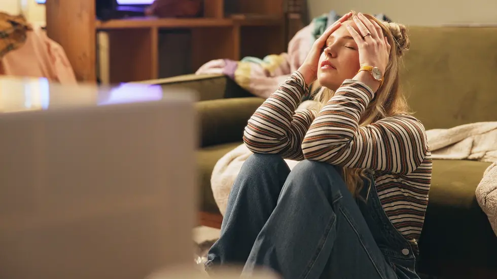 Woman in striped shirt and jeans sits on floor, head in hands, looking stressed. Sofa and colorful blanket in background, soft lighting.