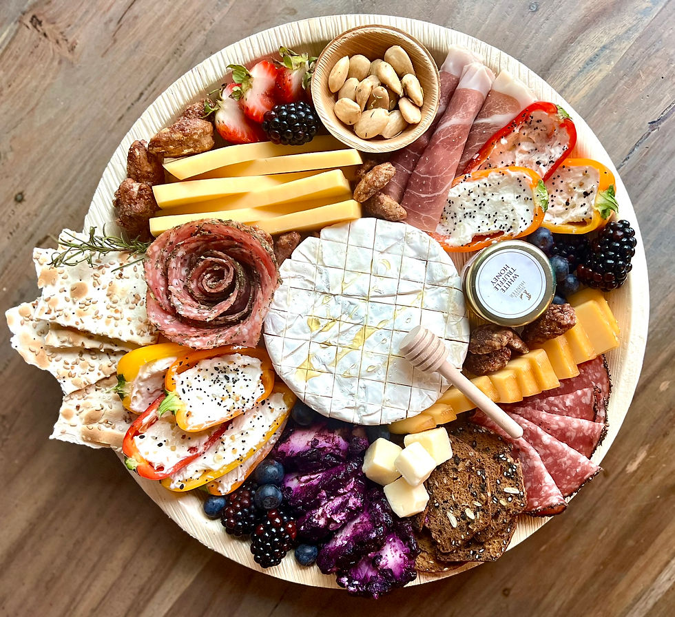 a round charcuterie board sits atop a wooden table and shows a scored cheese block, honey comb wooden stick, salami roses, cream cheese stuffed peppers and more