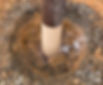 Wooden post partially wrapped and set in a gravel-filled hole, surrounded by orange-brown dirt. No text or people present.