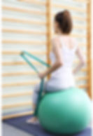 Frau, die auf einem Pezziball vor einer Sprossenwand sitzt und eine Therabandübung macht.