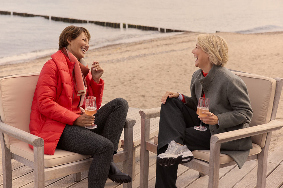Zwei Frauen sitzen gemeinsam am Strand, lachen und halten Getränke in der Hand.