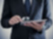 Man in suit examining tablet with a magnifying glass, suggesting scrutiny or investigation. Background is neutral and blurry.