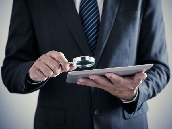 Man in suit examining tablet with a magnifying glass, suggesting scrutiny or investigation. Background is neutral and blurry.