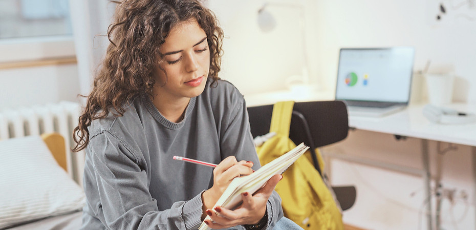 Student writing in a notebook with a laptop nearby, preparing for a college application essay.