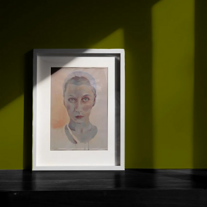Gerahmtes Porträt eines Gesichts mit hellen Augen in einem weißen Rahmen. Das Bild steht auf einem dunklen Boden vor einer grünen Wand.