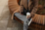 Eine Frau sitzt auf einer geschwungenen Holzbank im Freien, hält einen Kaffeebecher und schaut auf ihr Smartphone. Sie trägt einen beigen Mantel, graue Jeans und schwarze Converse-Sneaker.