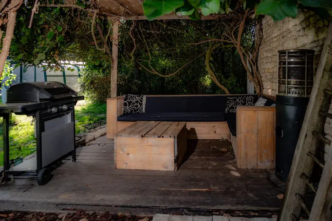 Overdekte zithoek op houten vloer onder klimplanten met houten bank, houten salontafel, barbecue links en staande terrasverwarmer rechts tegen stenen muur.
