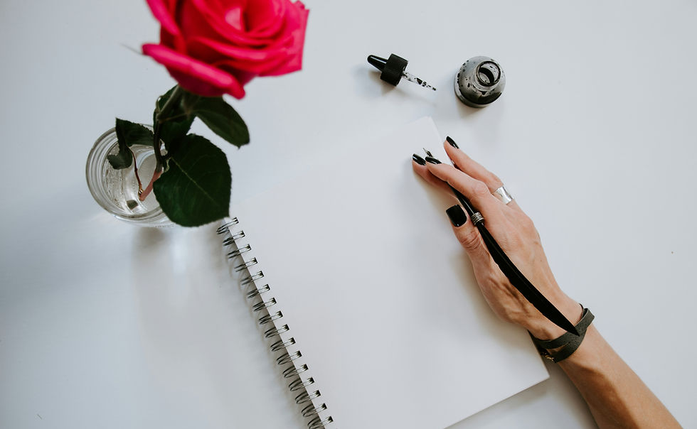 Hand with black nails holds a pen over a blank notebook. A red rose in a glass vase and ink bottle are on a white table, creating a creative mood.