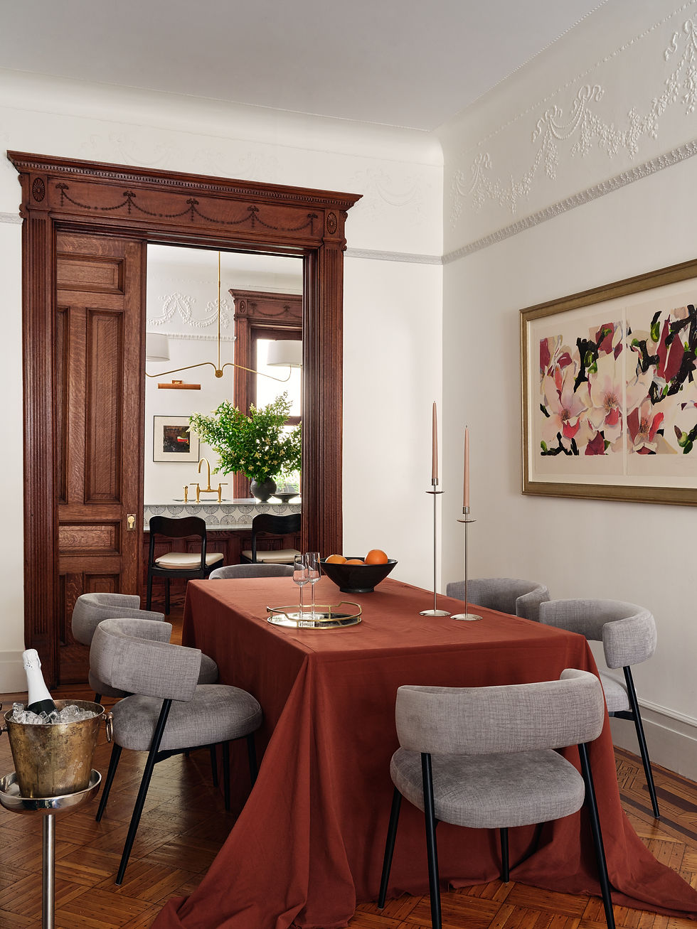 Elegant dining room with a rust-colored tablecloth draped to the floor, surrounded by soft gray upholstered chairs. A brass tray with glassware and two tall taper candles sits at the center of the table. The space features intricate white plaster molding, a large floral artwork, and a richly carved wood doorway that frames a view into a second room with a modern brass chandelier and greenery. A champagne bucket on a stand adds a festive, celebratory touch.