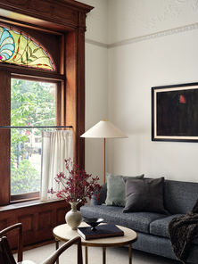 Historic Brooklyn brownstone living room with restored wood trim and stained glass window. Designed by Lex & Hudson, this sophisticated yet inviting space features a charcoal sofa with textured pillows, brass and marble coffee table, and sculptural floor lamp — blending modern comfort with original architectural details.