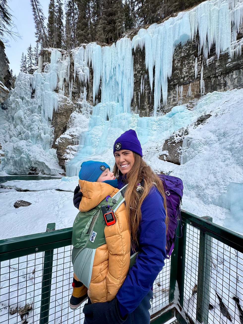 mom hiking to frozen waterfall with baby standing in front of falls with winter hiking buntings on.