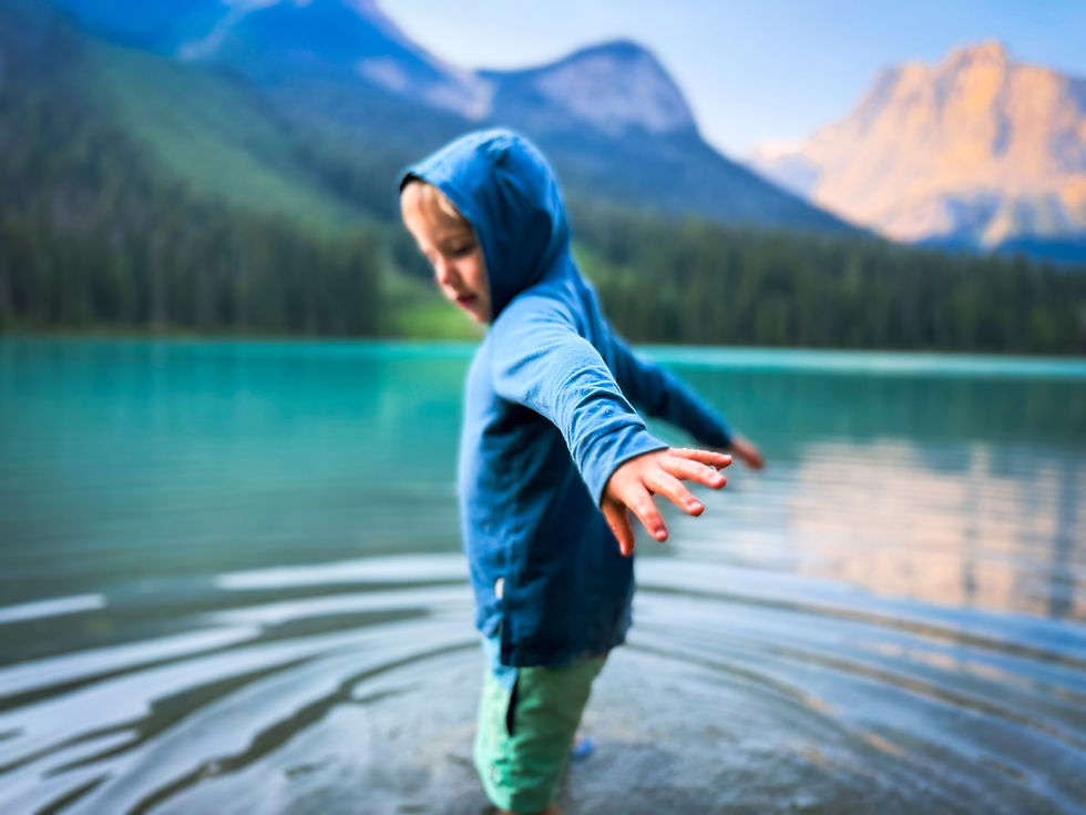 Young child in a blue hooded jacket stands in a turquoise mountain lake with arms out, calm and playful scene