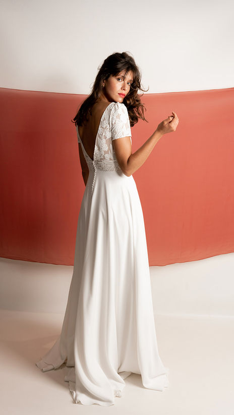 Robe de mariée en mousseline brodée florale avec un décolleté V froncé sous la poitrine et manches courtes, portée par une femme aux cheveux lâchés devant un fond rouge terracotta.