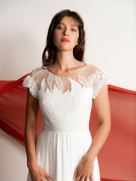 Femme portant une robe de mariée ivoire avec haut en tulle transparent orné de motifs floraux en broderie. Manches courtes volantées. Fond blanc avec drapé rouge vaporeux derrière elle.