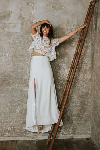 Femme souriante posant contre une échelle en bois, vêtue d’un top court en tulle brodé de fleurs et manches bouffantes et d’une jupe longue fendue sur le côté, sur un fond mural brut au style industriel.