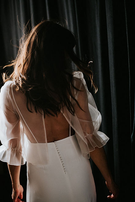 Vue de dos d'une femme portant un top transparent en organza de soie à manches longues bouffantes et une robe taille marquée boutonnée, dans une lumière tamisée devant des rideaux sombres.