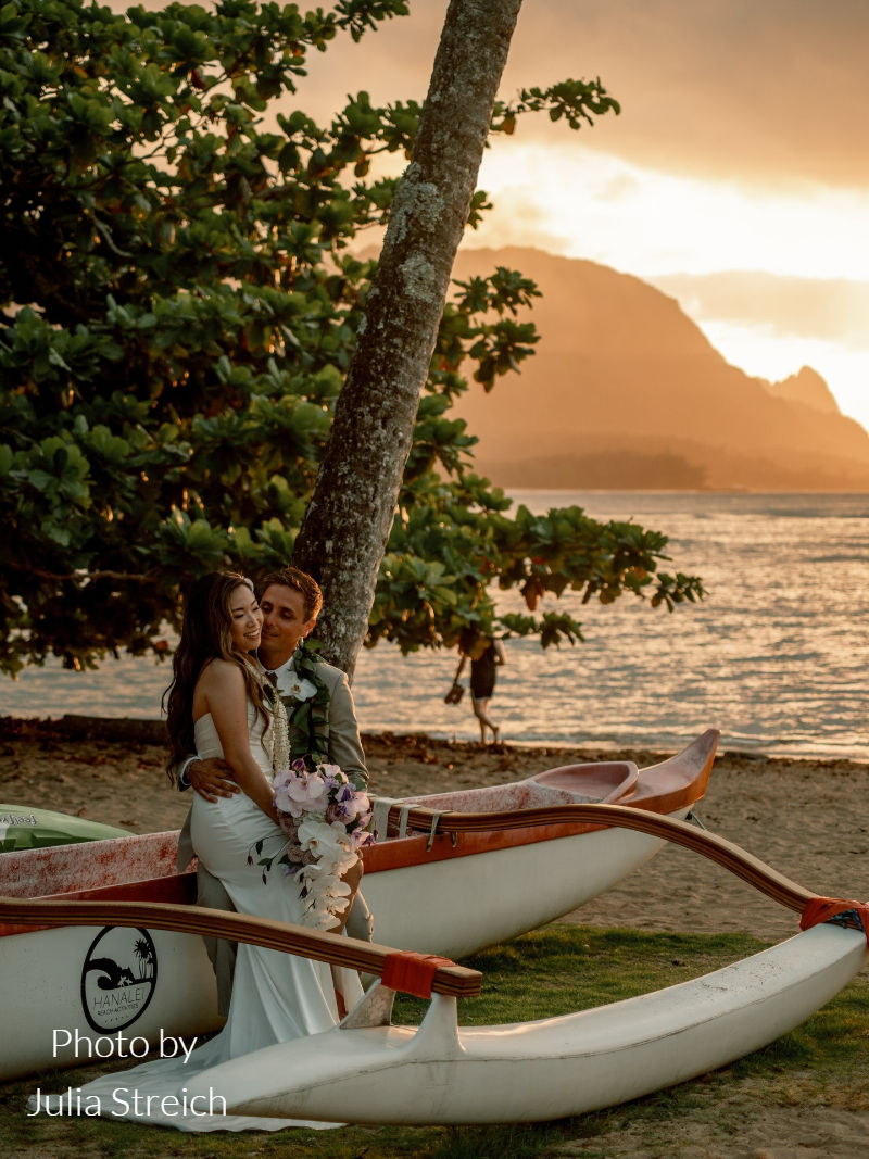 Couple embracing by canoe at sunset during destination wedding in Hawaii with ocean views and mountains