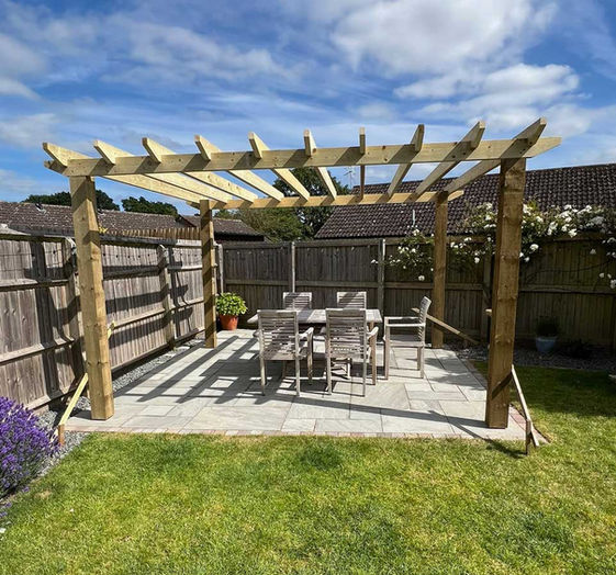Wooden pergola with table and chairs on a patio with landscaping.
