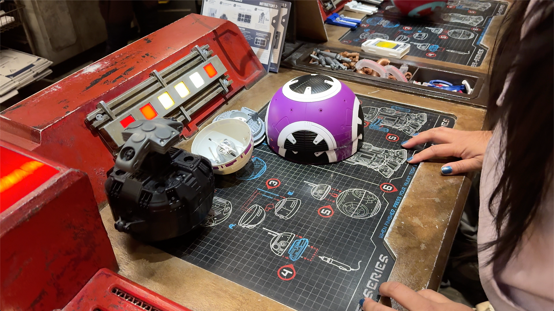 Chrissa assembling a BB series droid with the parts laid out in front of her with a diagram on the table.