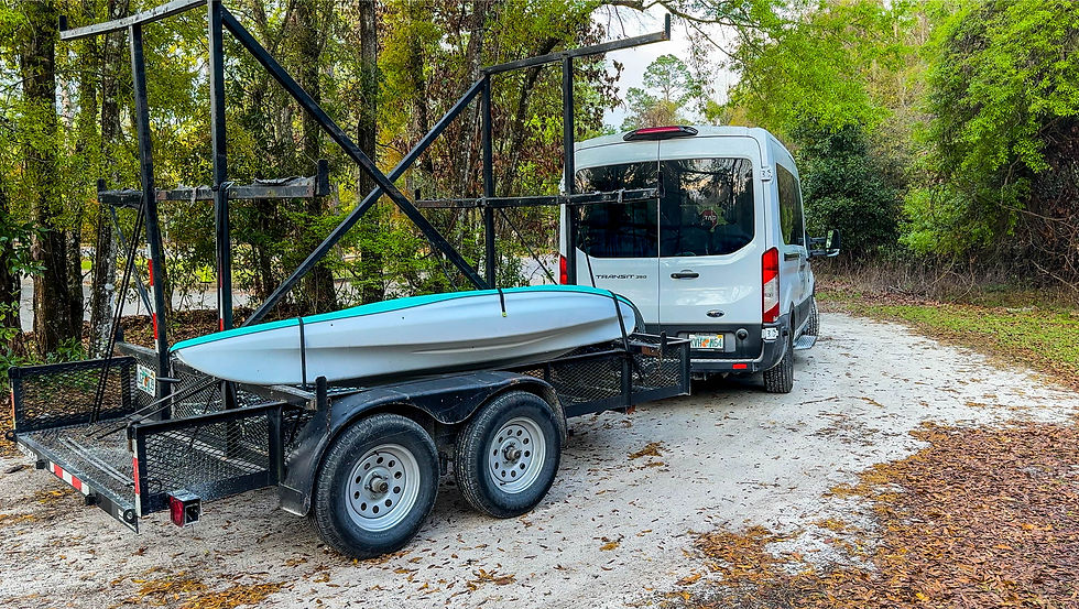 A white van and trailer with a kayak loaded on it surrounded by greenery