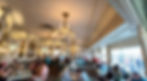 Elegant restaurant with diners at tables, ornate chandeliers, and mirrored walls. Bright atmosphere with people enjoying meals and conversations.