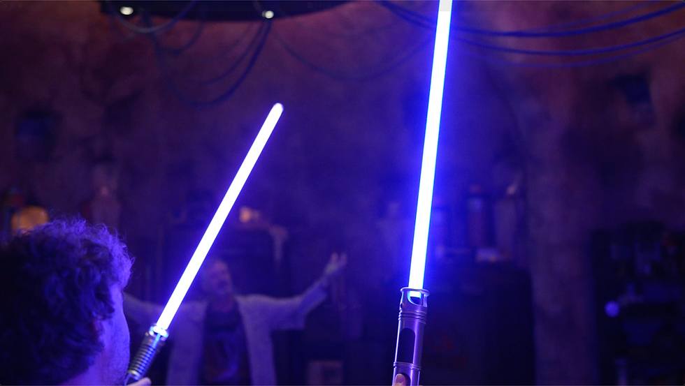 Two people hold glowing blue lightsabers in a dim room with a cave-like background. One person is blurred, raising their arms.