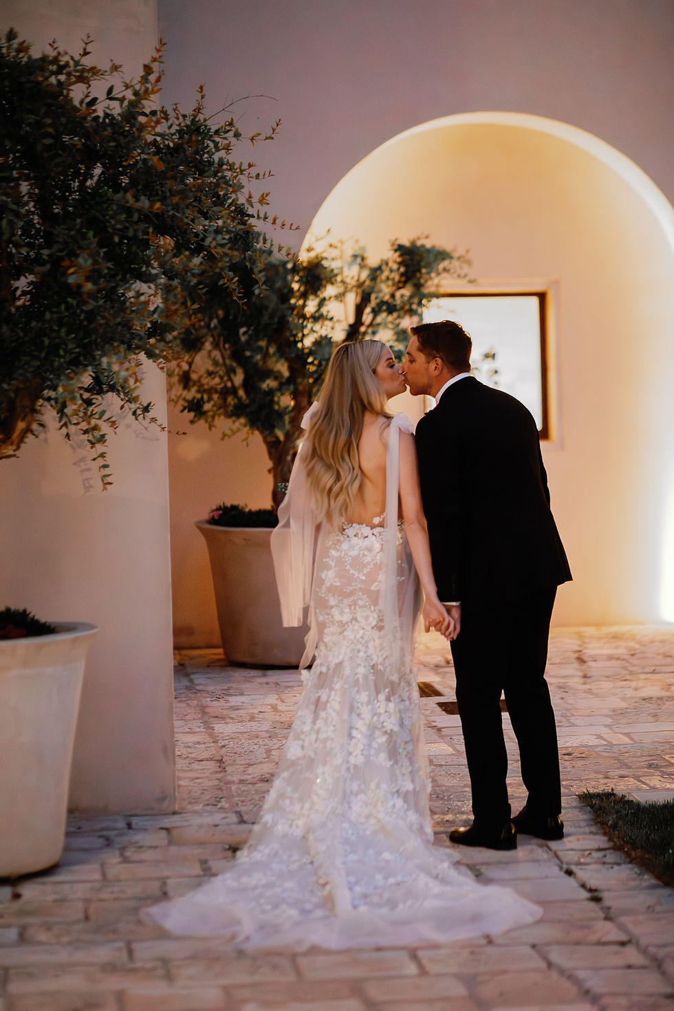 A couple kisses under an archway, she in a lace wedding dress, he in a black suit. Romantic setting with potted trees and warm lighting.
