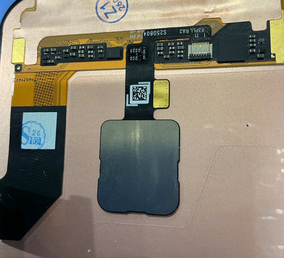 青い背景上に置かれたスマホ画面の内部部品の接写。金色の基板と黒いフレックスケーブル、銀色シールド、QRコードと数字印字が見える。