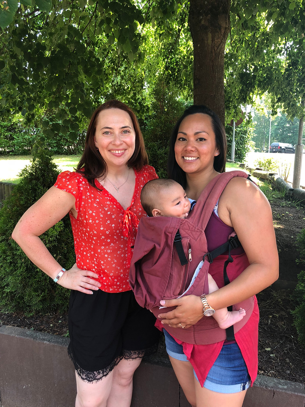 Two women standing in a park smiling. One woman is wearing a baby in a carrier