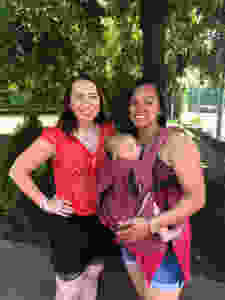 Two women standing in a park smiling. One woman is wearing a baby in a carrier