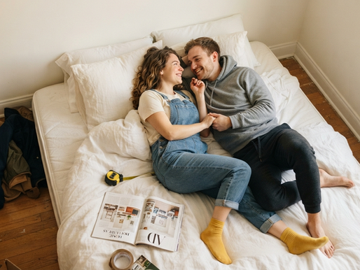 Casal sorrindo deitado na cama em quarto quase vazio, com caixas, fita métrica e revista, passando sensação de mudança.