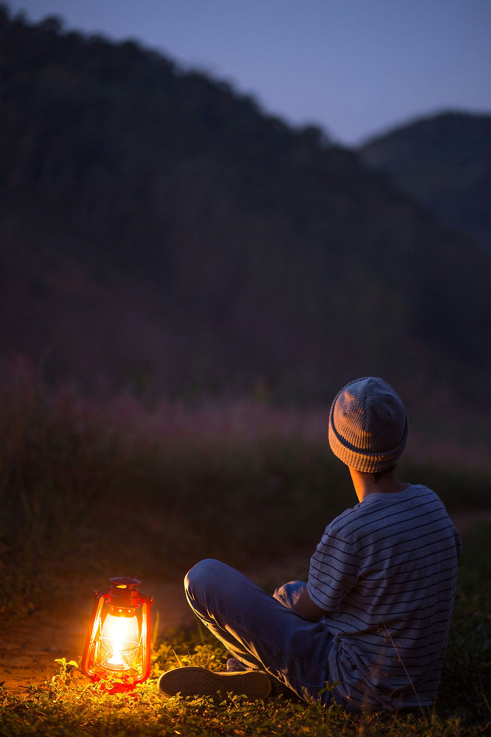 Person in striped shirt and beanie sits on grass at dusk, gazing at lantern. Mountains in the background. Warm, reflective mood.