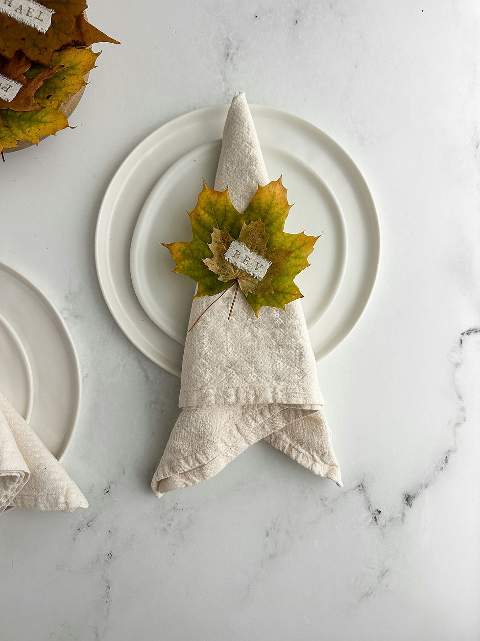 Beige napkin on white plate, topped with autumn leaves and a small tag reading REY, on a marble table.