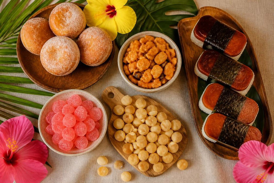 Overhead flat-lay of assorted Hawaiian snacks including malasadas dusted with cinnamon sugar, arare crackers, li hing mui gummies in bright pink, roasted macadamia nuts scattered on a wooden board, and spam musubi wrapped in nori, arranged with tropical hibiscus flowers and palm leaves on a natural linen background