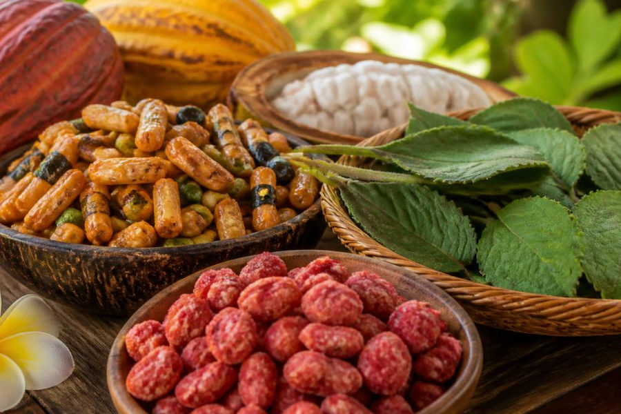Close-up of artisan Hawaiian snacks displayed on a wooden surface, including Li Hing Mui candies with their distinctive red coating, crispy arare crackers, and fresh island ingredients like cacao pods and mamaki leaves arranged nearby with natural island sunlight