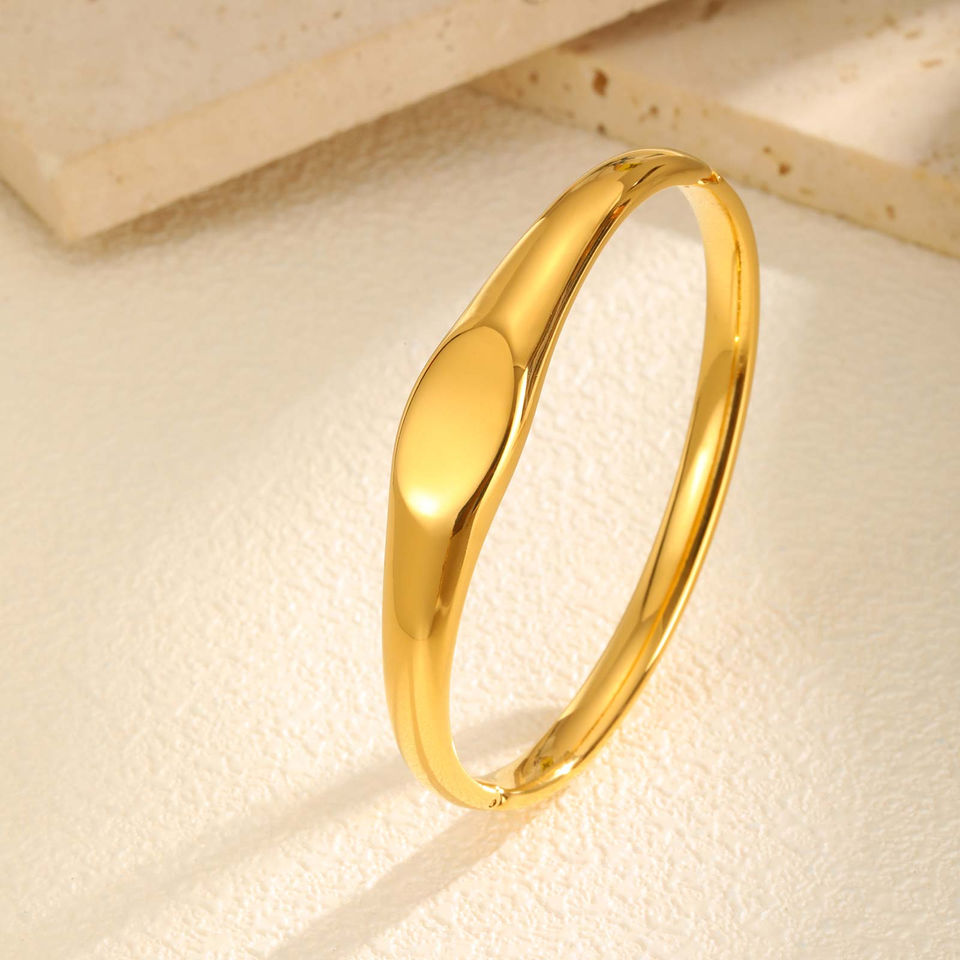 Elegant gold bangle bracelet, oval design, on beige surface. 9to5Jewels.
