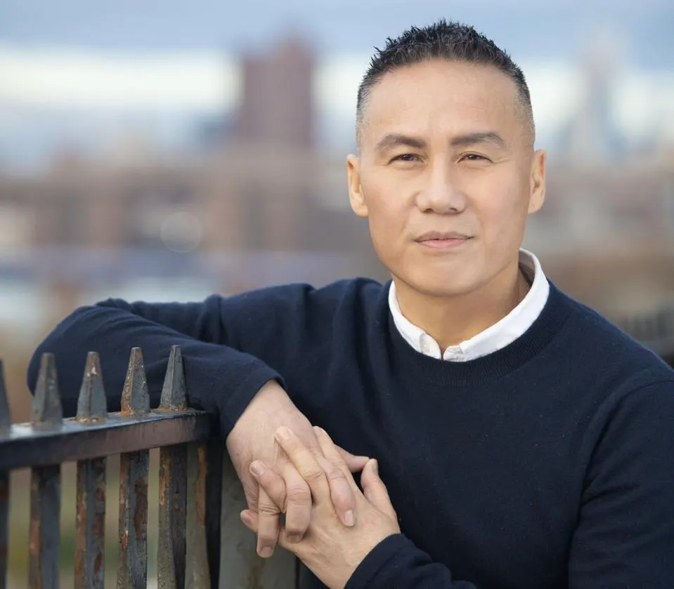 A portrait of BD Wong, wearing a black sweater, and posing with his hands together. 