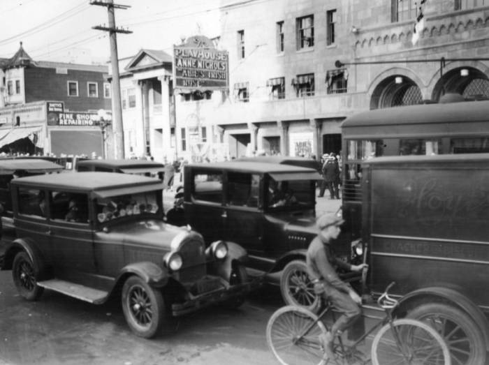 A black and white image of the front of the theater, behind various cars from the 1920's and a boy riding a bicycle